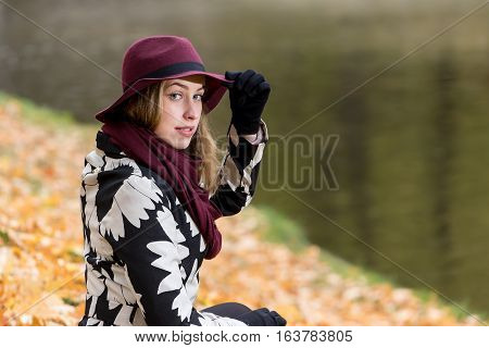 Woman in a floral patterned coat and wine red hat in the park by the river. Happy girl and colorful autumn forest. Portrait of a lady yellow green red and brown leaves background