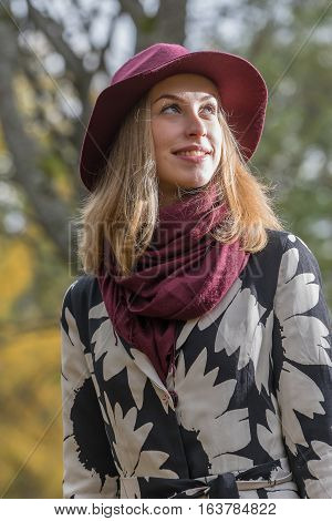 Woman in a floral patterned coat and wine red hat in the park. Happy girl and colorful autumn forest. Portrait of a lady yellow green red and brown leaves background