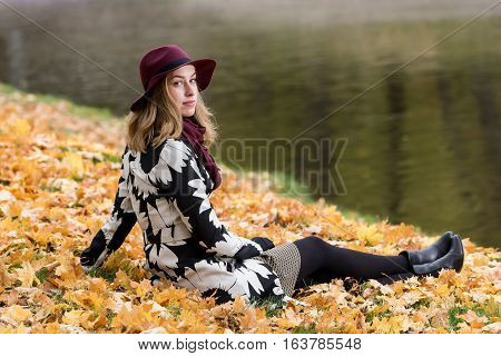 Woman in a floral patterned coat and wine red hat in the park by the river. Happy girl and colorful autumn forest. Portrait of a lady yellow green red and brown leaves background