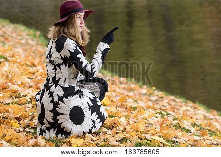 Woman in a floral patterned coat and wine red hat in the park by the river. Happy girl and colorful autumn forest. Portrait of a lady yellow green red and brown leaves background