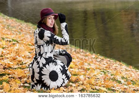 Woman in a floral patterned coat and wine red hat in the park by the river. Happy girl and colorful autumn forest. Portrait of a lady yellow green red and brown leaves background