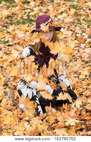 Woman in a floral patterned coat and wine red hat playing with leaves in the park. Happy girl throws maple tree leaves into the air. Yellow green red and brown leaves colorful autumn forest