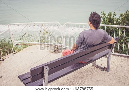 Young Man Sitting On Bench Overlooking Sea