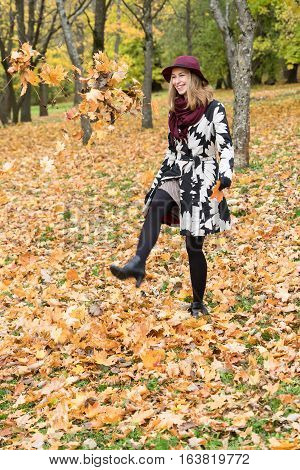 Woman in a floral patterned coat and wine red hat playing with leaves in the park. Happy girl throws maple tree leaves into the air. Yellow green red and brown leaves colorful autumn forest