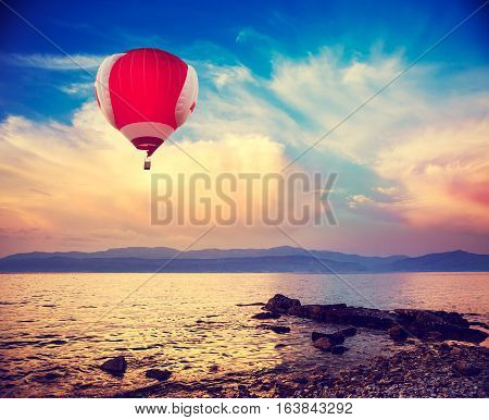 Hot Red Air Balloon Flying over Sea at Sunset. Blue Sky Background with Beautiful Clouds. Pebble Beach and Evening Seascape. Coastline and Mountains on the Horizon. Toned and Filtered Photo.
