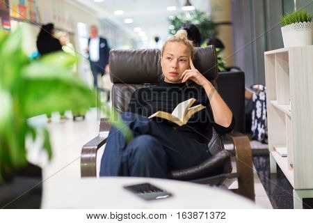 Thougtful woman sitting by wooden table and reading book in public library.