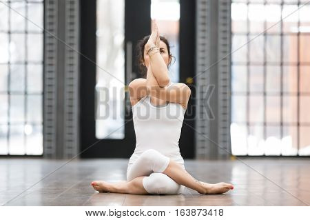 Full length portrait of attractive young woman working out in luxury fitness center, doing yoga or pilates exercise without mat on wooden floor. Gomukasana, Cow Face pose