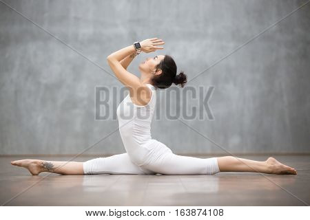 Attractive young woman wearing white sportswear set and smartwatch working out against grey wall, doing yoga or pilates exercise. Monkey god, splits, hanumanasana. Full length