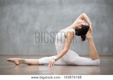 Young pretty woman wearing white sportswear set and smartwatch working out against grey wall, doing yoga or pilates exercise. Variation of monkey god, splits, hanumanasana with backbend. Full length