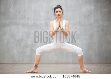 Portrait of beautiful young woman wearing white sportswear working out against grey wall, doing yoga or pilates exercise. Standing in Sumo Squat, Goddess pose. Full length