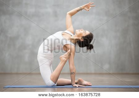 Side view portrait of beautiful slim young woman with tattoo working out in fitness center against grey background, doing yoga or pilates exercise. Variation of ustrasana, camel pose. Full length