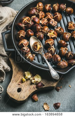 Roasted chestnuts in cast iron grilling pan over rustic wooden board and grey concrete stone background, selective focus, vertical composition