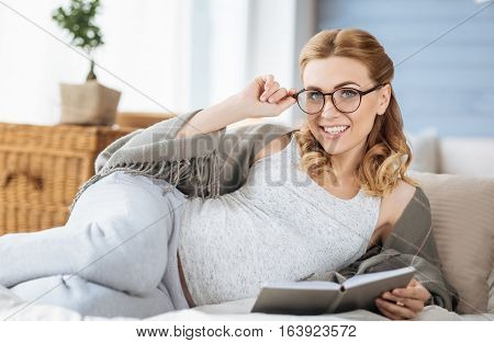 Enjoy leisure time. Pregnant young overjoyed woman reading book in bed while smiling and posing.