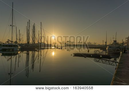 panoramic view sunrise at the Valencia harbor, the sun rises between docked sailboats and cargo port cranes, water reflection