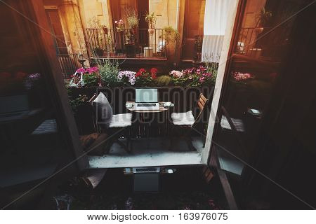 Small cozy beautiful workspace on balcony with laptop mouse smartphone chairs and flowers around on sunny summer day seeing through opened doors from dark interior facade of another house behind