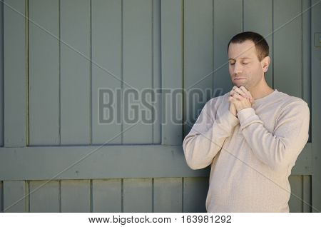 Man praying outdoors in front of a green wooden background.