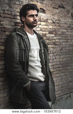 Young male urban city style on wall. A beautiful young man wearing winter clothes in the street. Pants, white sweater and green long coat. Hands in his pockets, leaning on an old wall. Intense attractive look, stylish hair.