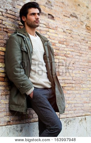 Young male fashion street on wall. A beautiful young man wearing winter clothes in the street. Pants, white sweater and green long coat. Hands in his pockets, leaning on an old wall. Intense attractive look, stylish hair.