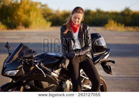 Outdoor Shot Of Pleased Female Bikes Puts On Leather Gloves, Dressed In Black Clothes, Poses On Moto