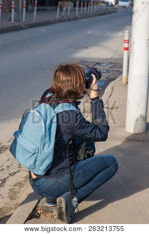 Photographer In The Street Photo Shooting Process. Model Is Young Lady In Stylish Black Coat With Bl