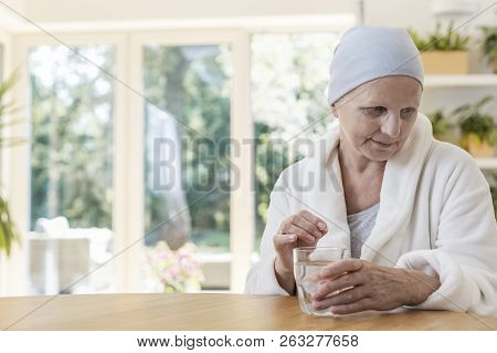 Senior Woman Suffering From Ovarian Cancer Wearing Bathrobe And Headscarf Taking Pills At Home.