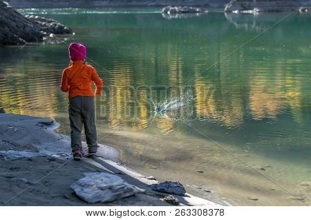 A Little Girl, A Child, In Warm Clothes And A Red Cap Throws Stones Into The Water From The Sandy Sh