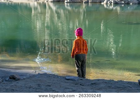 A Little Girl, A Child, In Warm Clothes And A Red Cap Throws Stones Into The Water From The Sandy Sh