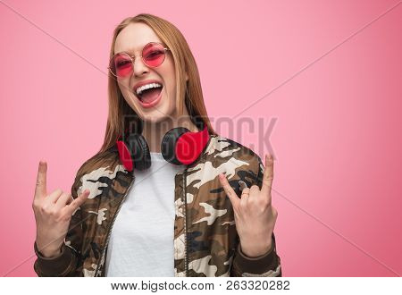 Trendy Blond Woman In Colorful Sunglasses And Jacket With Earphones Showing Rock And Roll Gesture Wh