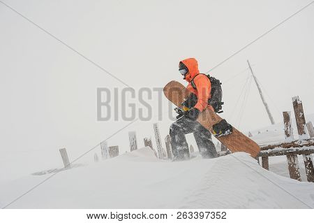 Man Walking With The Snowboard In Hands In The Mountain Up The Hill Near The Fence In The Popular To