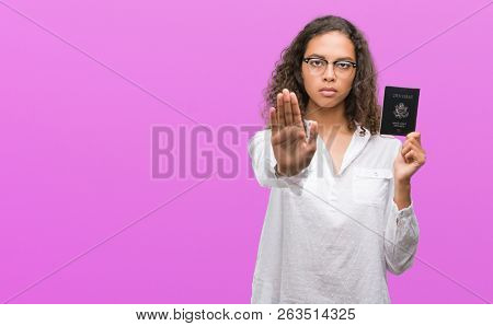 Young hispanic woman holding passport of United States of America with open hand doing stop sign with serious and confident expression, defense gesture