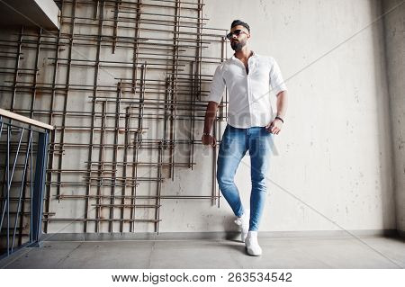 Stylish Tall Arabian Man Model In White Shirt, Jeans And Sunglasses Posed Against Steel Wall Indoor.