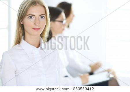 Attractive Businesswoman At Meeting Or Conference Against The Background Of Colleagues. Group Of Bus