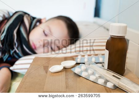 Young Boy Wearing Sweater And A Scarf Sleeping In His Bed While Having A Flu. Meds And Syrips On A W