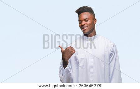 Young arabic african man wearing traditional djellaba over isolated background smiling with happy face looking and pointing to the side with thumb up.