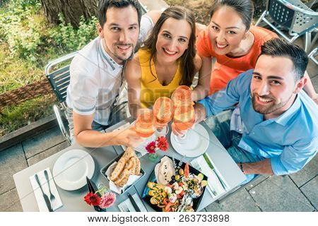 High-angle view portrait of four cheerful and beautiful young friends, looking at camera while toasting at a trendy restaurant with delicious food outdoors in summer