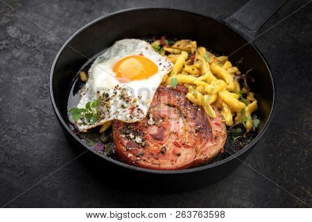 Traditional fried Allgauer veal roast roll sausage slices with fried egg and spaetzle in a cast-iron skillet with spices as closeup on a black board 