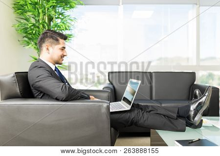 Full Length Of Relaxed Male Professional Using Laptop While Sitting In Office Lobby