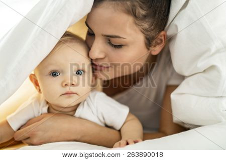 Portrait Of Young Mother With Infant Baby Girl Liying On The Bed Covered With A White Blanket