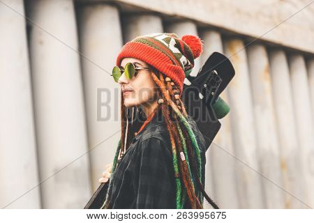 Young Girl In Sun Glasses And A Cap Stands With A Longboard Against The Background Of Concrete Struc