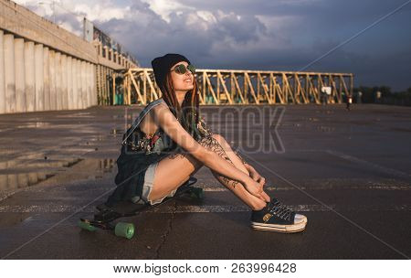 Young Girl With Tattoos And Dreadlocks In A Blue Cap Sits On A Longboard Against The Background Of C