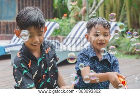 Cute Boys Are Blowing A Soap Bubbles Outside. Indonesian Or Malasian Brother Siblings Are Playing To