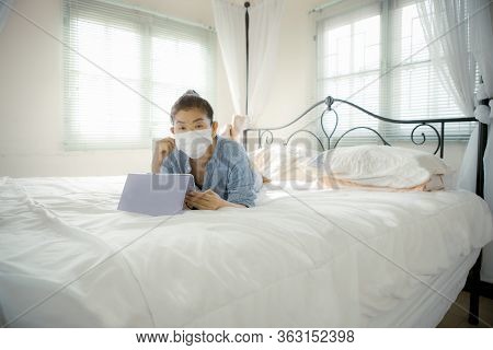 Woman With Protection Mask In Hand Lying On Sofa Bed In Home Living Room Chatting To Internet Device