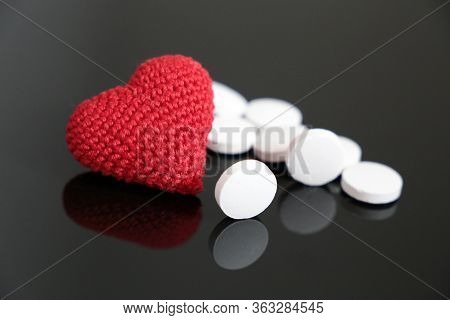 White Pills And Knitted Red Heart On Dark Glass Table. Concept Of Hypertension, Heart Disease, Cardi