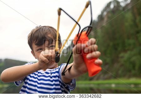 Young Guy Takes Aim, Preparing To Make Shot From Slingshot. Portrait Boy Is Going To Shoot On Backgr