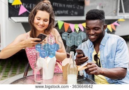 eating, technology and people concept - happy mixed race couple with smartphones and takeaway chinese wok and drinks at food truck over nutritional value chart