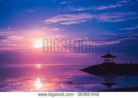 Pink Dawn The Sun And The Gazebo At Sanur Beach, Bali, Indonesia. A Calm Surface Of Sea Water That R