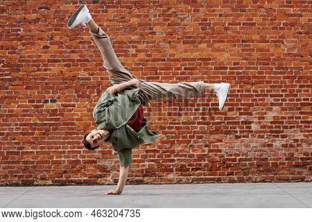 Full Length Shot Of Young Man Doing Hip-hop Handstand Pose And Smiling At Camera Against Brick Wall,