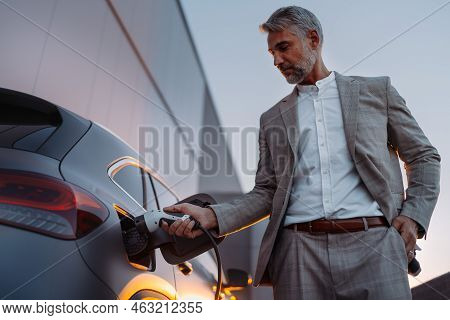 Businessman, Holding Power Supply And Charging His Electric Car During Sunset. Concept Of Ecology Tr