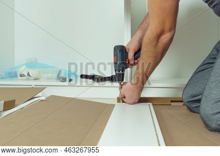 Male Hands Hold Cordless Electric Screwdriver, Screw To Fix Walls Of White Drawer, Assembling Dresse