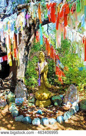 October 5, 2022 In Mt Shasta, Ca:  Tibetan Prayer Flags Surrounding A Guanyin Sculpture Besides Tree